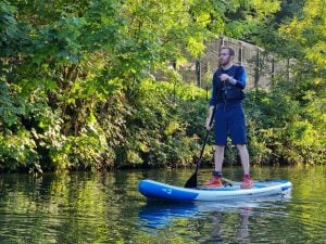 A man standing in shorts and t-shirt, wearing a life vest, paddling on a stand up paddleboard down a tree-lined river