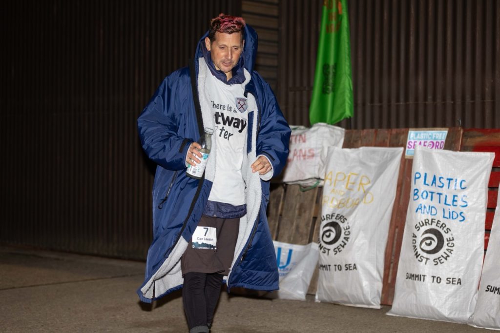 A runner wearing a thick coat and holding a water bottle passes a wall of recycling bags at a race. It is nighttime and the runner looks tired.