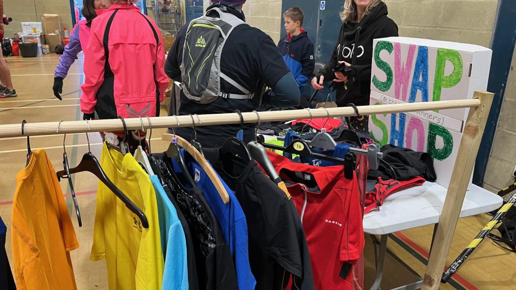 A community hall with runners speaking to volunteers. In the foreground is a clothes rail with running kit available to swap.