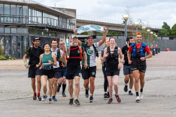 A group of runners jogging together with one holding a torch and another a flag with the green runners' logo on it. All are wearing a Green Runners badge.