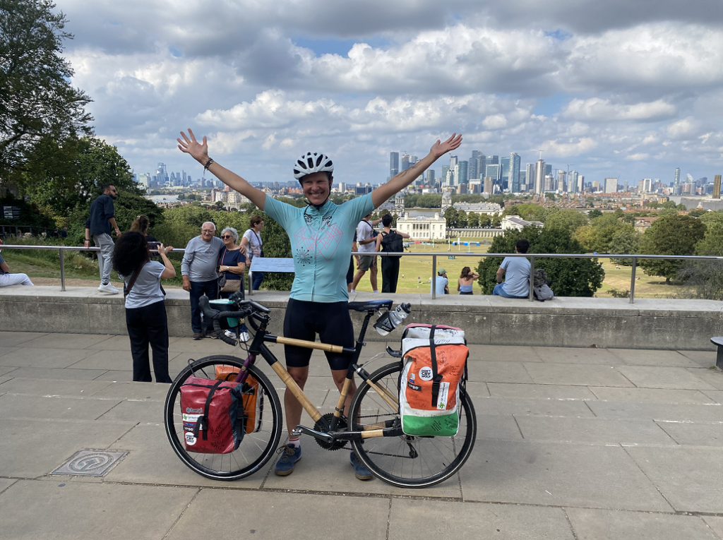 Kate Strong standing behind her bamboo bike with arms raised, and London skyline in the background.