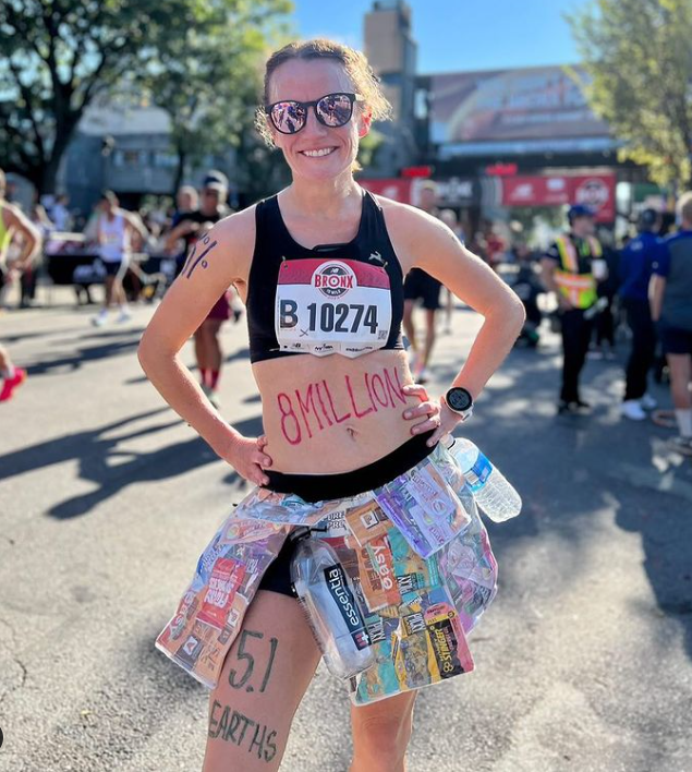 Marathon runner Tina Muir poses hands on hips wearing a skirt made from upcycled waste plastic and with "8 million" written on her tummy and "5.1 earths" written on her right leg.