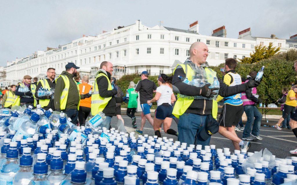 Brighton Marathon volunteers stand next to a huge table of plastic water bottles handing them out to passing runners.