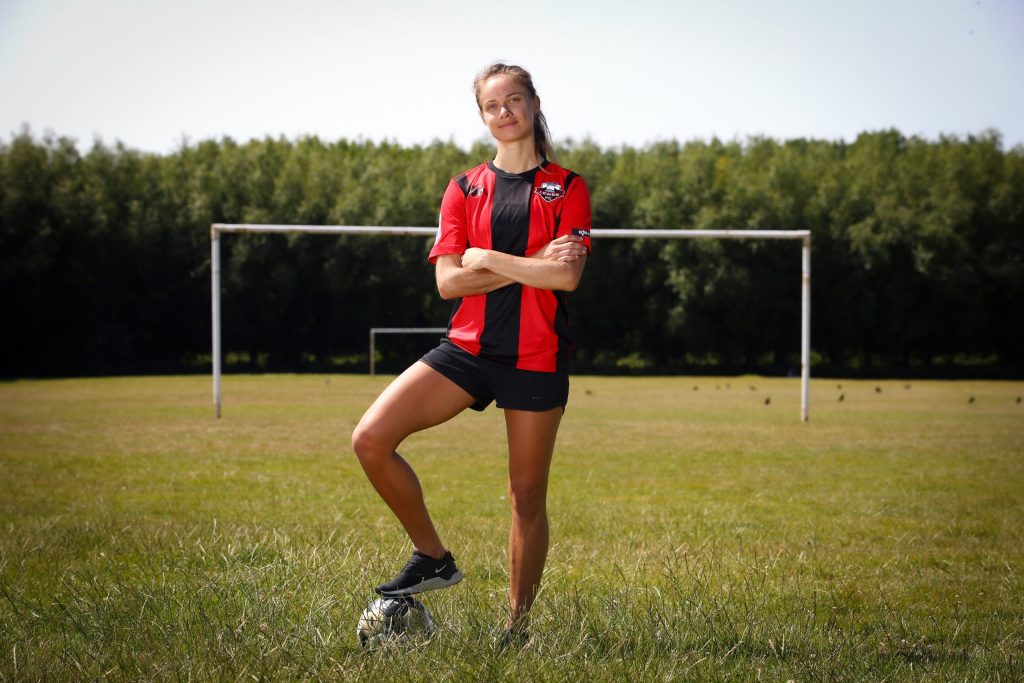 Katie Rood stands on a football pitch arms crossed wearing a red and black shirt with one foot on a football.