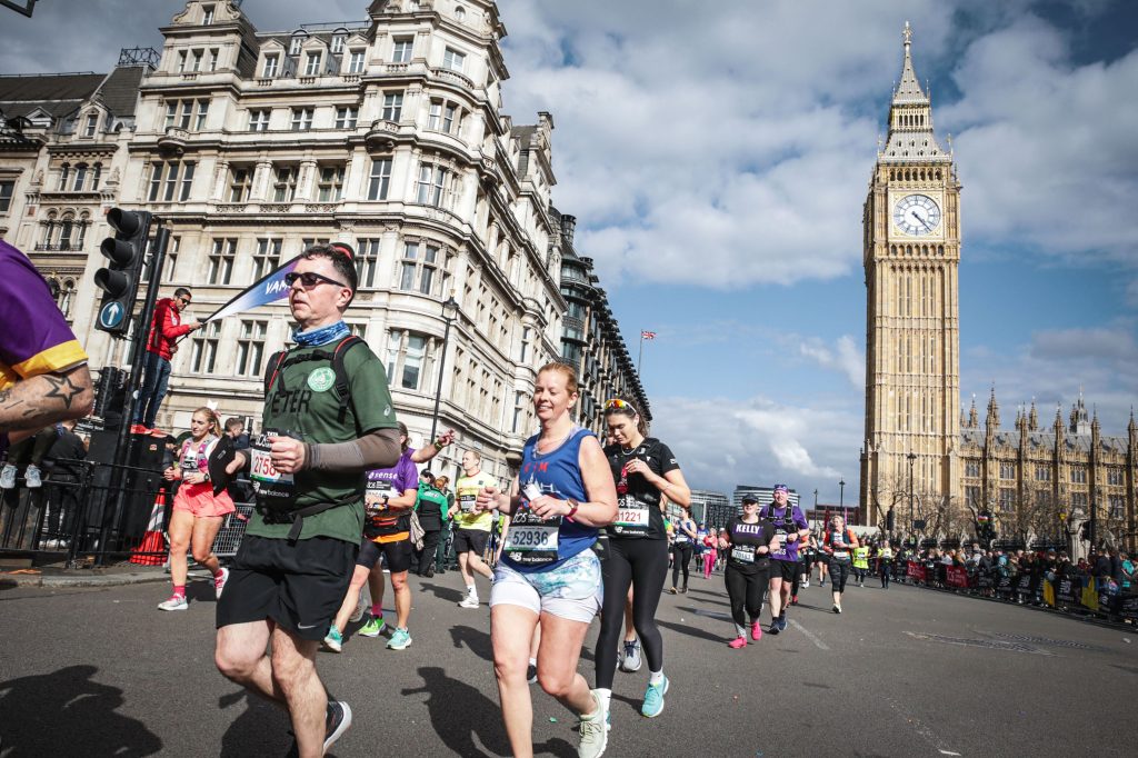 A photo of people running a marathon. London's Big Ben is in the background. There are lots of people in the picture and a person waving a banner. It is a sunny day.