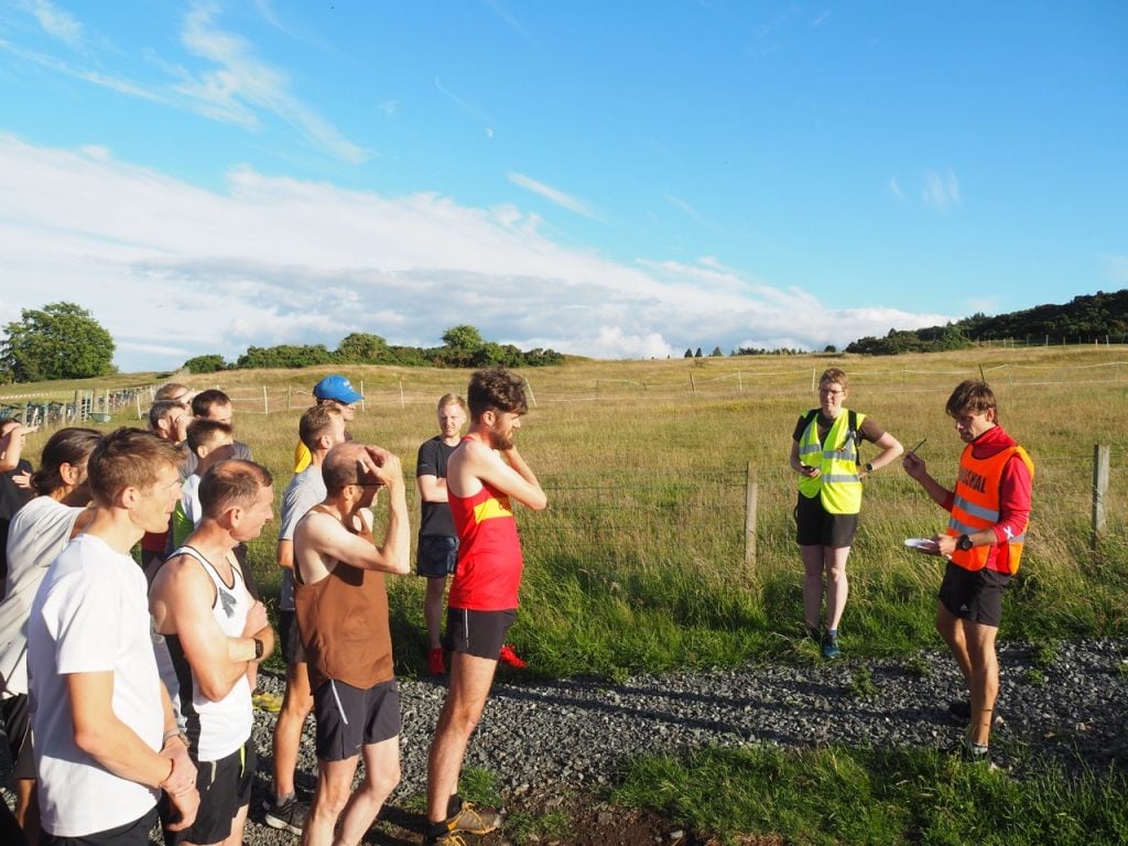 A group of runners on a start line with the race director at the front giving a briefing. It is a sunny evening.