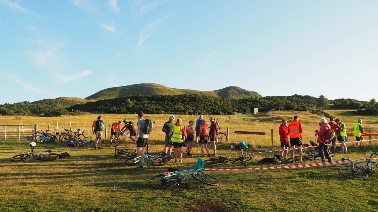 A sunset scene with grassy hills in the background framed against a blue sky. In the foreground, people standing surrounded by bikes talk at the start of a race
