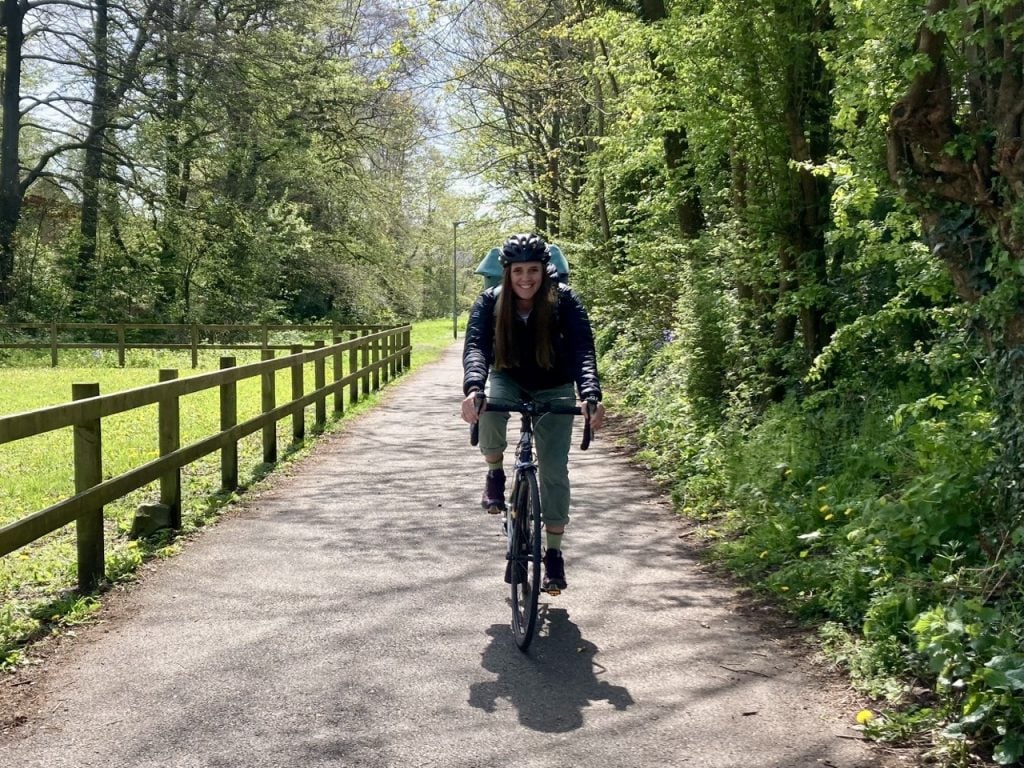 A woman cycling on a bike path towards the camera. She is smiling and it is a sunny day