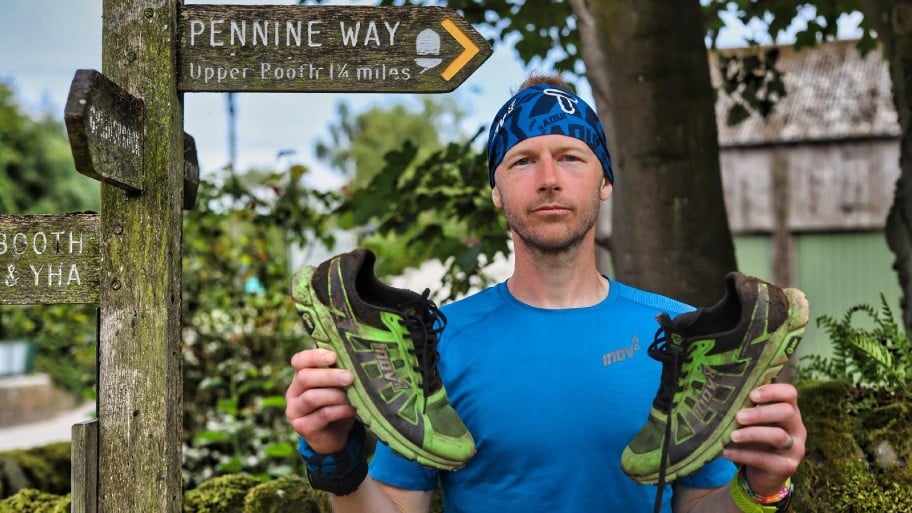 A man with a dodgy haircut holding two muddy running shoes beneath a sign that says 'Pennine Way'. He is wearing a blue top and a blue bandana.