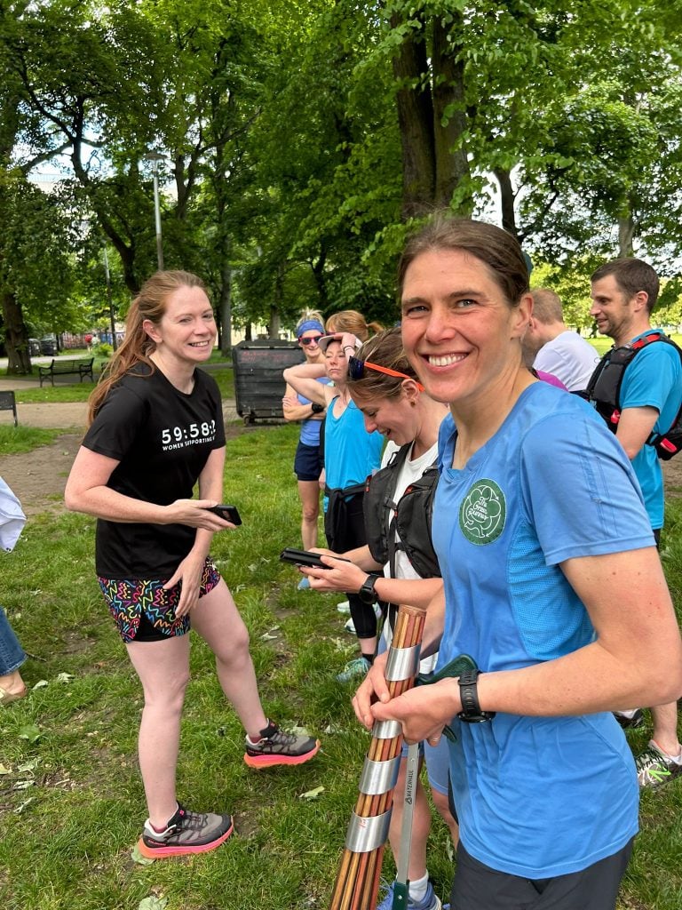 Jasmin Paris smiling in a leafy park holding a baton about to start her leg of the Climate Relay. She is slim with brown hair and she has other people behind her.
