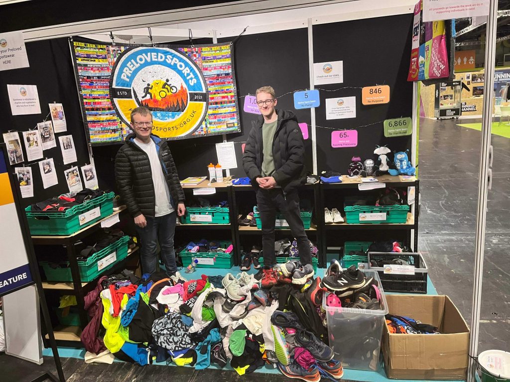 Two people wearing puffy jackets standing in a 3x2m square event tent. The tent is full of supermarket crates packed with donated running gear. At their feet is a pile of donated kit. Behind them is a Preloved Sports banner.