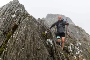 Kev climbs down a steep, rocky mountain face. He is wearing a grey jacket, black shorts, and a bib with the number "26." He is holding trekking poles for balance. The mountain peaks are shrouded in mist.