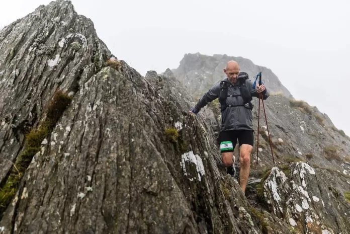 Kev climbs down a steep, rocky mountain face. He is wearing a grey jacket, black shorts, and a bib with the number "26." He is holding trekking poles for balance. The mountain peaks are shrouded in mist.