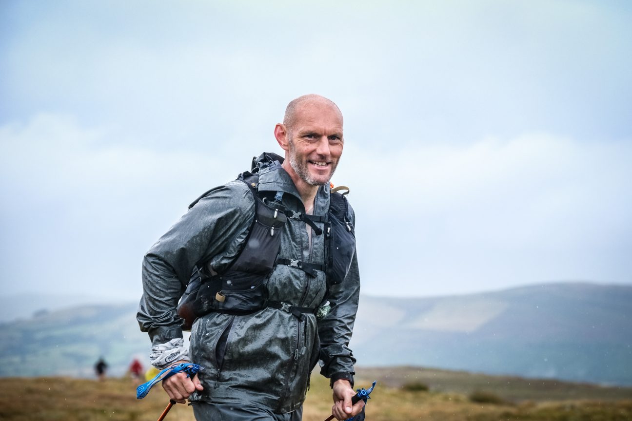 Kev holding his custom trekking poles, hikes through a field with rolling hills in the background under a cloudy sky.