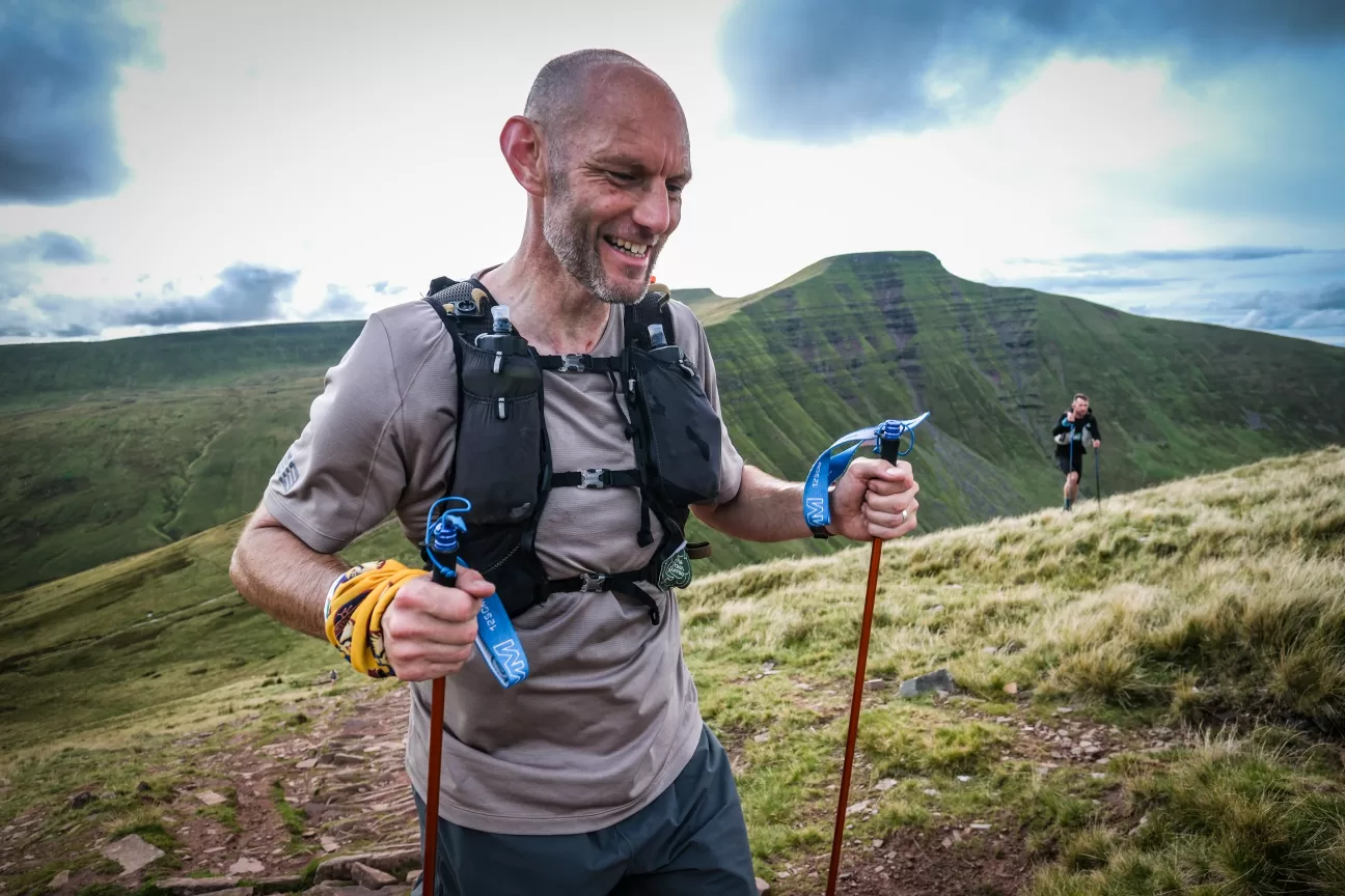 Kev smiles at the camera as he walks up a grassy hill. He is wearing a grey running shirt, a hydration vest, and shorts. He holds two trekking poles, and a yellow buff is visible on his left wrist. Another runner is in the background. The sky is cloudy, and mountains are visible behind him
